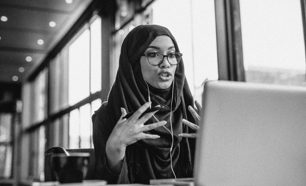 Businesswoman in hijab having a video chat on laptop while sitting at coffee shop. Female sitting at cafe and making video call using earphones and laptop computer.