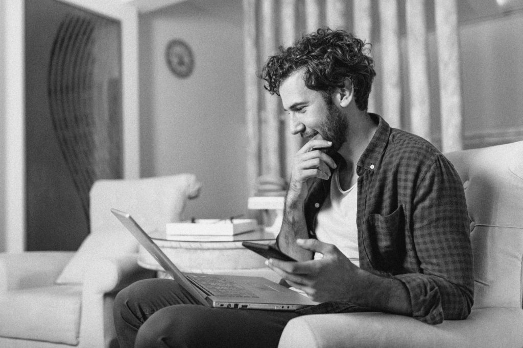 Successful entrepreneur smiling in satisfaction as he checks information on his laptop computer while working in a home office. Young man relaxing on the sofa with a laptop and mobile phone in hands