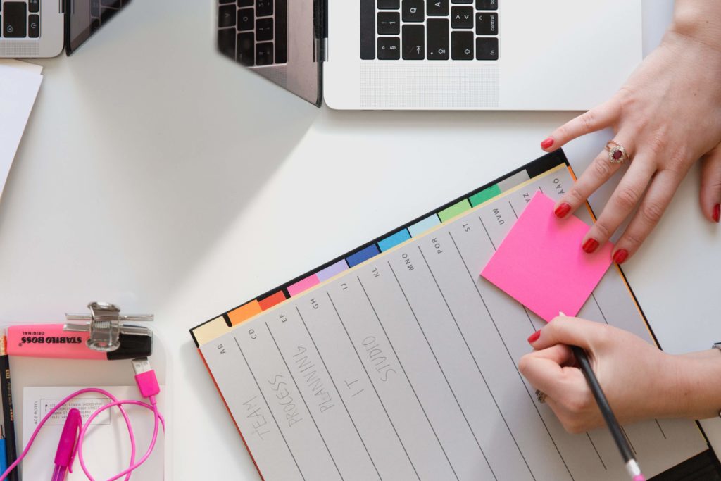 A person sits in front of a laptop and notebook, writing something on a pink sticky note.