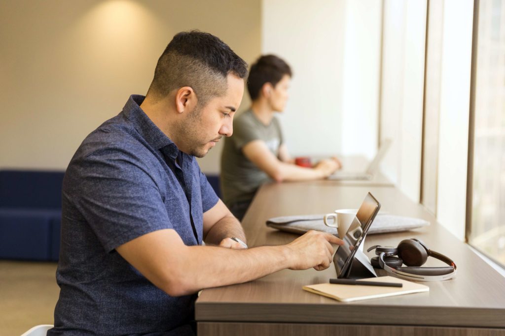 A man sits at a desk in front of a tablet. Headphones are also on the table in front of him. He is wearing a purple shirt.