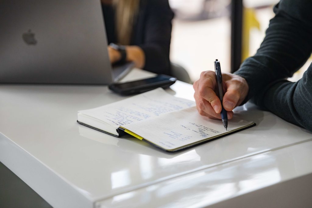 A person's hand is shown up close. They hold a pen in front of an open notebook.