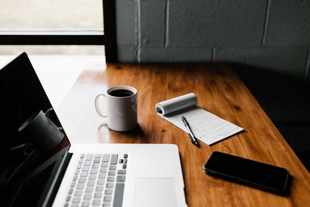 An open laptop is shown with a mug of coffee and a notebook to the right of it.