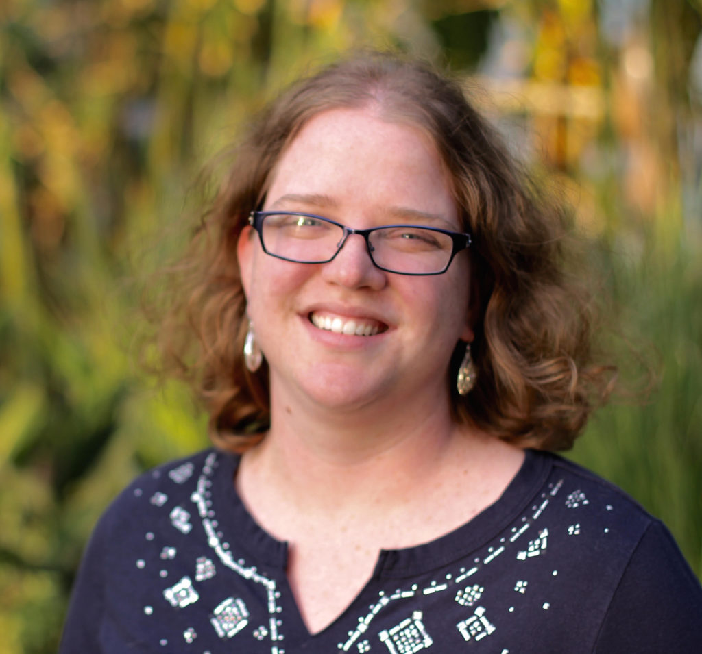 Carrie Miller is shown smiling at the screen. She wears a navy blue top and is standing against a green background.