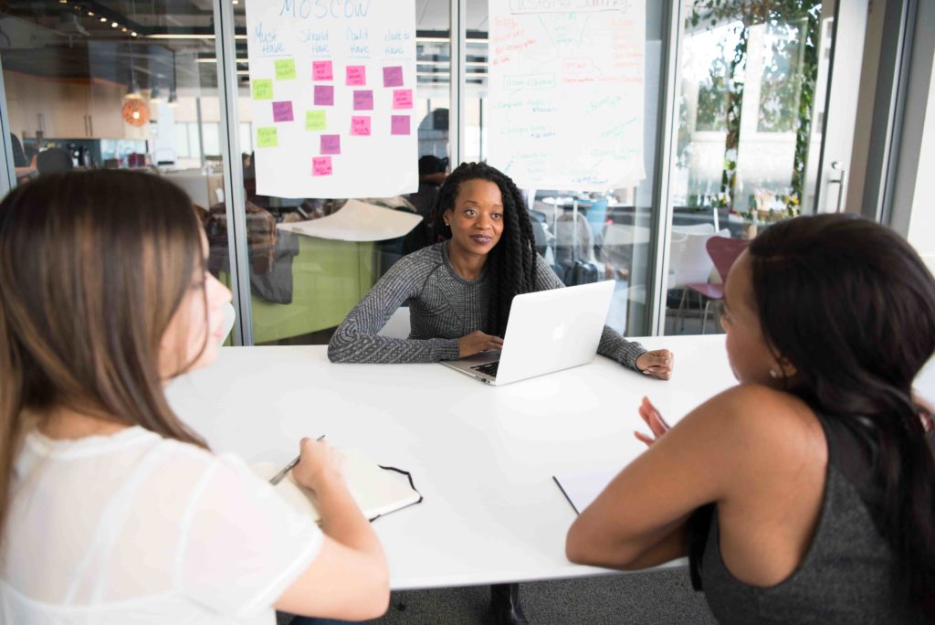 Three women sit at a table together. A page with sticky notes is attached to the wall behind them.